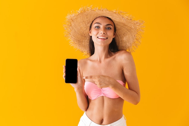 Jovem atraente e alegre usando biquíni isolado na parede amarela, mostrando tela em branco do celular