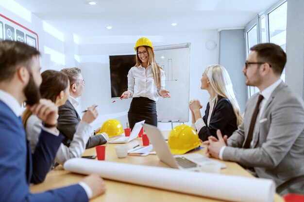 Jovem atraente e bem sucedida arquiteta caucasiana feminina, vestida com roupa formal e com capacete amarelo na cabeça, em frente ao quadro branco e falando. Seus colegas sentados e ouvindo.