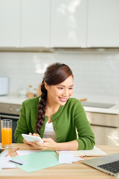 Jovem atraente e sorridente mulher pagando contas online usando laptop de casa enquanto está sentada na cozinha