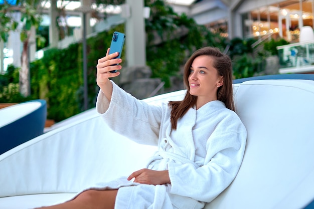 Jovem atraente e sorridente, vestindo um roupão branco, tirando uma foto de retrato de selfie na câmera do smartphone enquanto relaxa no spa resort