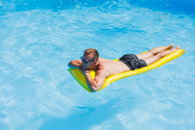 Jovem atraente em óculos de sol e shorts está relaxando em um colchão amarelo inflável na piscina Férias de verão