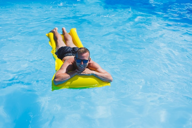 Jovem atraente em óculos de sol e shorts está relaxando em um colchão amarelo inflável na piscina férias de verão