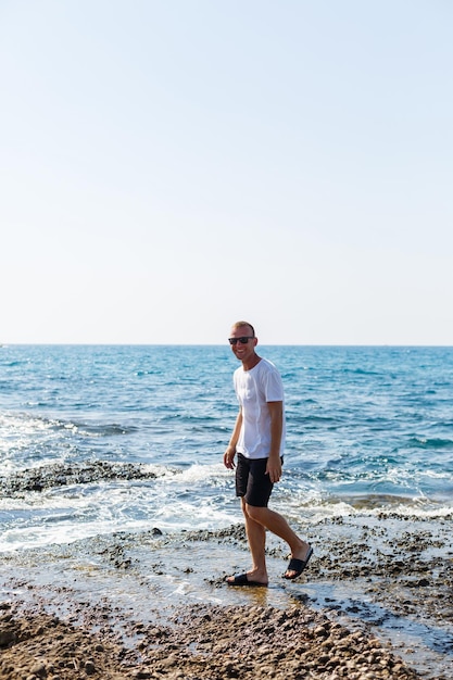 Jovem atraente em óculos de sol em uma camiseta branca e shorts fica na costa do mar mediterrâneo