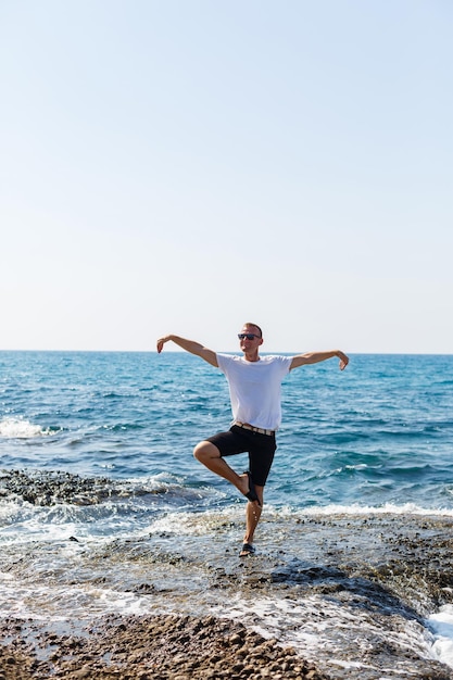 Jovem atraente em óculos de sol em uma camiseta branca e shorts fica na costa do mar mediterrâneo