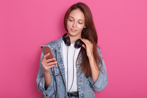 Jovem atraente em pé isolado sobre fundo rosa, segurando o smartphone na mão, com fones de ouvido no pescoço e toca com os dedos, olha para o dispositivo, vestindo roupa elegante