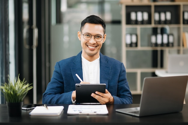 Jovem atraente empresário asiático sorrindo pensando planejando escrever em notebook tablet e laptop olhando para a câmera no escritório