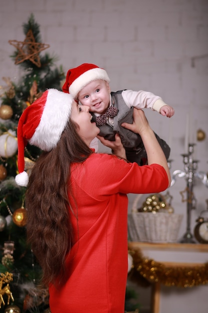 Foto jovem atraente feliz mãe com chapéu de papai noel se divertir com seu filho em casa perto da árvore de natal. família, felicidade, feriados, conceito de ano novo