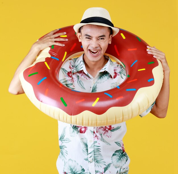 Jovem atraente homem asiático com chapéu e camisa havaiana branca com padrões de flores vermelhas e verdes e anel de natação donut em volta do pescoço, indo para as férias na praia contra um fundo amarelo.