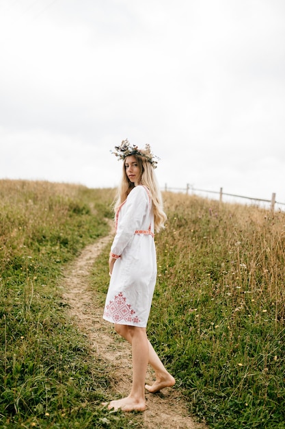 Jovem atraente loira com os pés descalços em um vestido branco com ornamento e guirlanda de flores na cabeça posando no campo