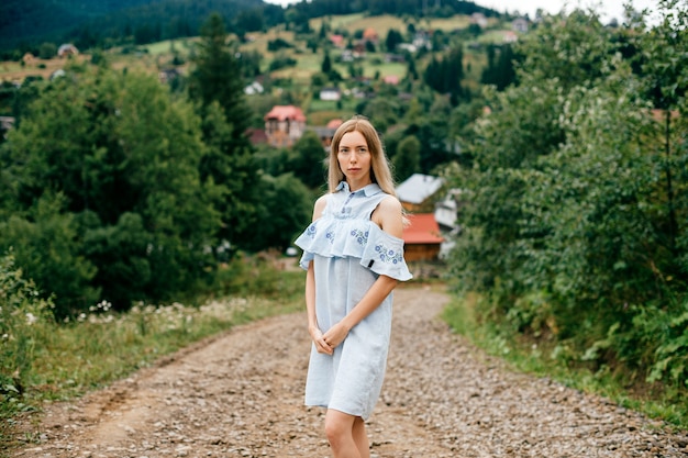 Jovem atraente loira elegante com vestido azul romântico, posando na estrada na zona rural