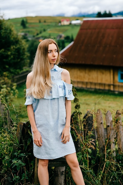 Jovem atraente loira elegante em um vestido azul posando sobre uma casa de campo no campo