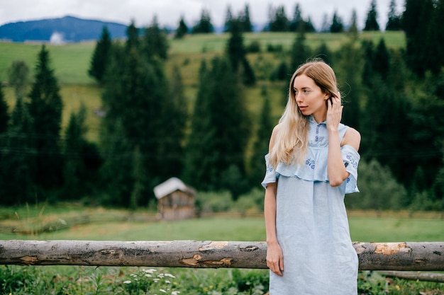 Jovem atraente loira elegante em um vestido azul romântico posando perto de uma cerca de madeira no campo