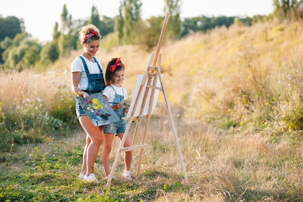 Jovem atraente mãe ensina filha a pintar no parque de verão. conceito de atividade ao ar livre para crianças em idade escolar