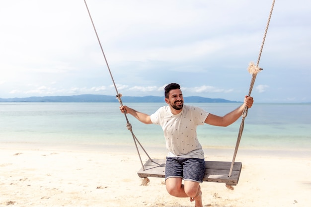 Jovem atraente mistura feliz correu homem balançando em um balanço na costa de um mar tropical brilhante