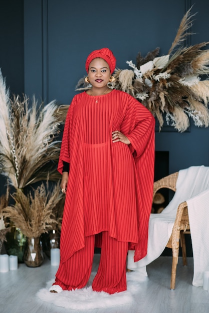 Jovem atraente modelo feminino africano em um elegante terno vermelho e lenço na cabeça, posando no estúdio com decorações rústicas boho no fundo. Foto de corpo inteiro de uma senhora africana elegante