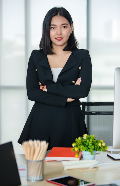 Jovem atraente mulher asiática em terno preto de pé, braço cruzado com confiança em um escritório moderno. Conceito de estilo de vida de escritório moderno.