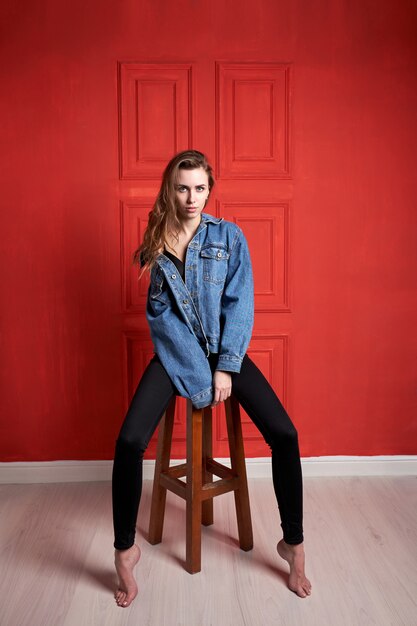 Jovem atraente ou modelo com cabelos longos, vestido com uma jaqueta jeans e calça preta, localização na cadeira.