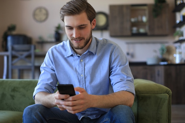 Jovem atraente relaxando em um sofá em casa e usando o telefone celular para cheking redes sociais.