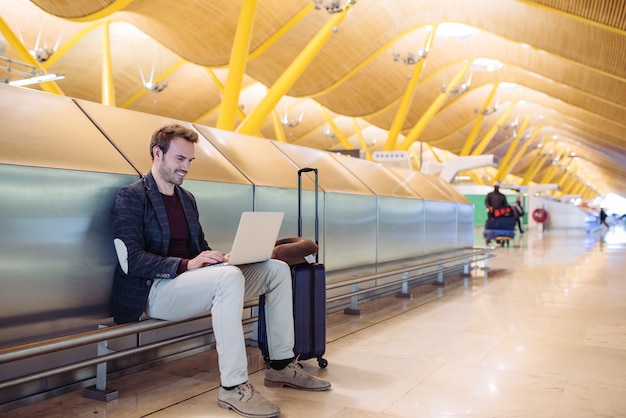 Jovem atraente, sentado no aeroporto, trabalhando com um laptop, esperando seu voo com uma mala