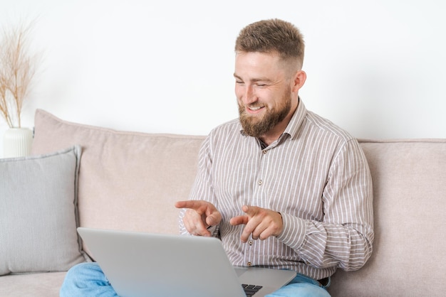 Jovem atraente sentado no sofá em casa trabalhando no laptop online usando