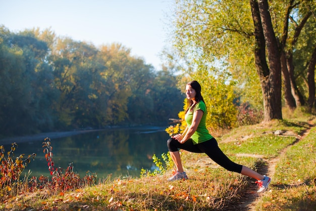 Jovem atraente sorridente esportes mulher estendendo-se enquanto a manhã corre na floresta perto do rio. Sentindo-se bem, relaxado, feliz. Conceito de estilo de vida saudável