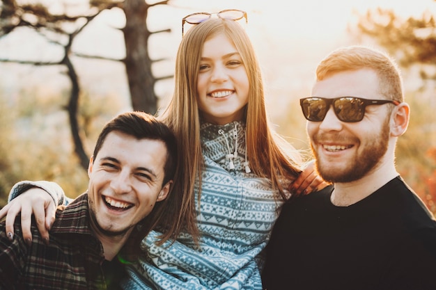 Jovem atraente sorrindo e abraçando dois homens bonitos, enquanto passam um tempo na natureza em um dia ensolarado. Mulher abraçando os homens na natureza