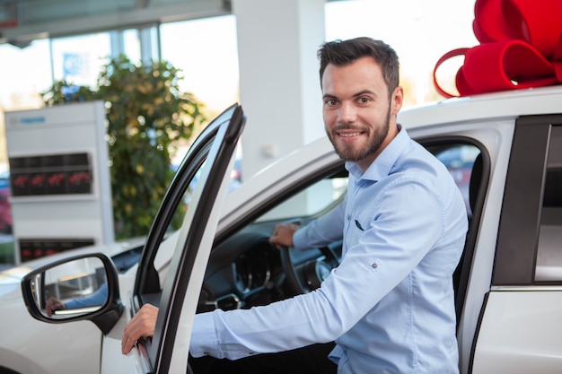 Jovem atraente, sorrindo para a câmera, enquanto entra em um automóvel novo para venda na concessionária