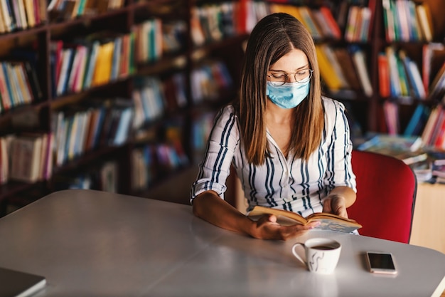 Jovem atraente universitária com máscara facial, sentado na biblioteca e estudando durante a pandemia de corona.