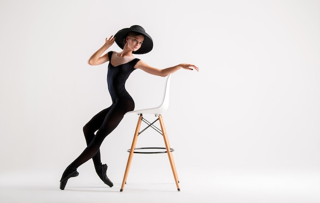 Jovem bailarina com sapatilhas pretas e chapéu elegante posando sentada na cadeira isolada no fundo branco