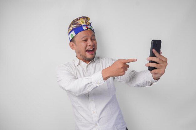 Jovem balinês de camisa branca e chapéu tradicional fazendo uma videochamada com família ou amigo, sentindo-se muito feliz e entusiasmado