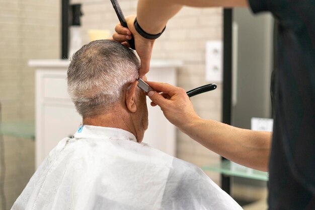 Jovem barbeiro usando a navalha para terminar um corte de cabelo de velho