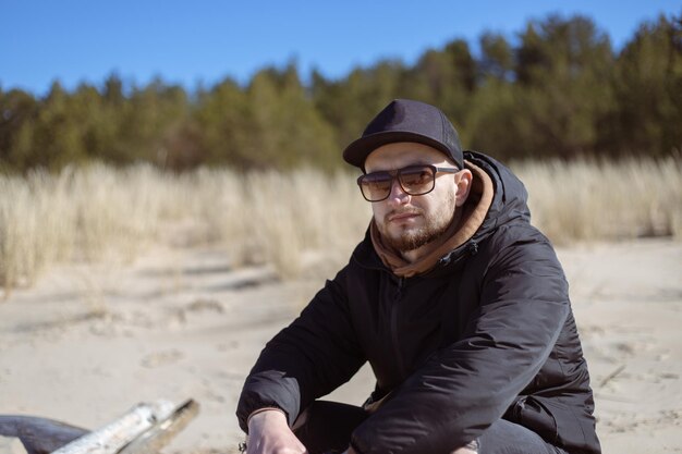 Jovem barbudo caucasiano sentado em um tronco na praia de areia