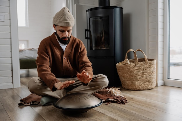 jovem barbudo com um instrumento musical melodioso que emite sons cósmicos glucophone ou handpan foto elegante de tocar tongdrum no interior de uma casa de madeira branca