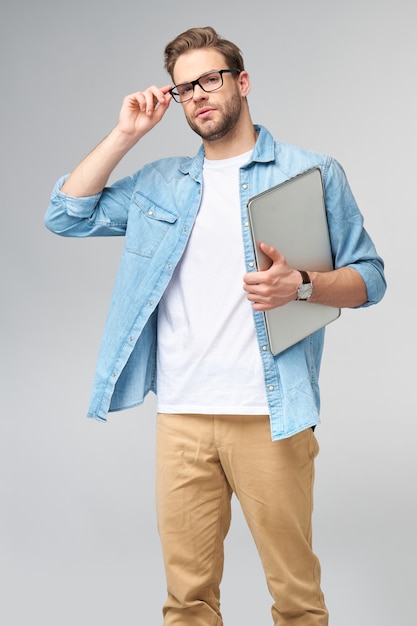 Jovem barbudo concentrado de óculos, vestido com uma camisa jeans, segurando o laptop isolado