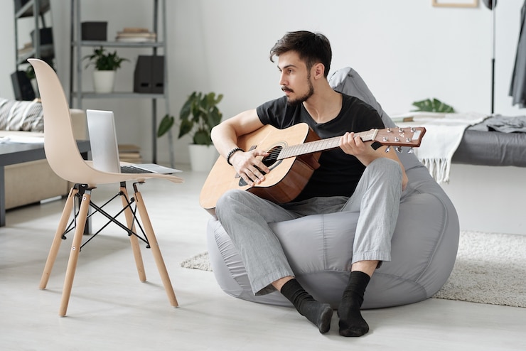 Foto jovem barbudo concentrado sentado em um saco de feijão e assistindo a um vídeo no laptop enquanto aprende a tocar guitarra