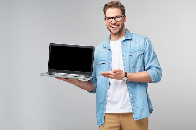 Jovem barbudo concentrado usando óculos e uma camisa jeans segurando um laptop