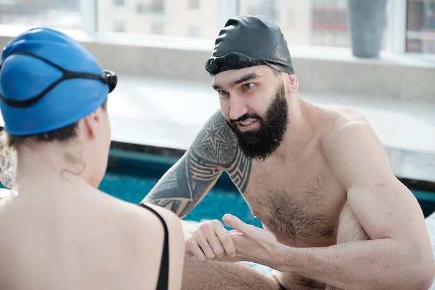 Jovem barbudo contente de touca de natação conversando com a nadadora enquanto torce por ela à beira da piscina