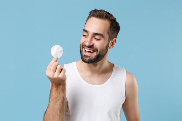 Jovem barbudo de 20 anos de idade em camisa branca segura almofada de algodão na mão isolada em fundo de parede pastel azul, retrato de estúdio. Conceito de procedimentos cosméticos de saúde de cuidados com a pele. Simule o espaço da cópia.