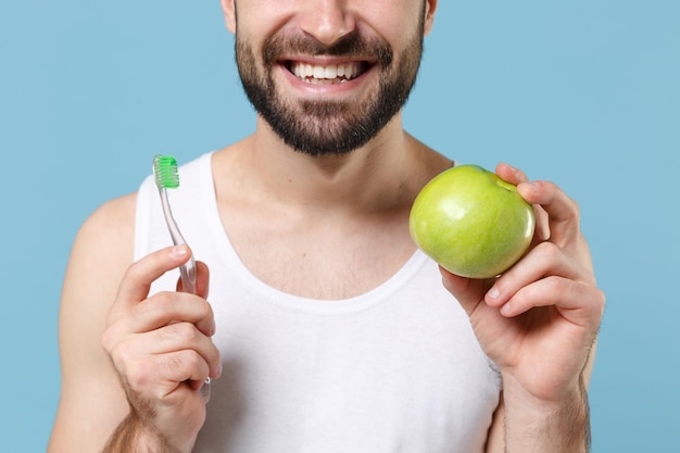 Jovem barbudo de 20 anos de idade em camisa branca segura pincel e maçã verde isolada em retrato de estúdio de fundo de parede pastel azul. Conceito de procedimentos cosméticos de saúde de cuidados com a pele Simule o espaço da cópia.