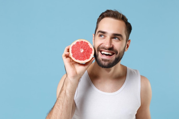 Jovem barbudo de 20 anos de idade na camisa branca segura na mão metade da toranja isolada em fundo pastel azul, retrato de estúdio. Conceito de procedimentos cosméticos de saúde de cuidados com a pele. Simule o espaço da cópia