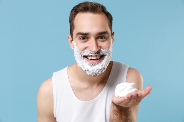 Jovem barbudo de 20 anos de idade no rosto de camisa branca coberto com espuma de barbear isolada em retrato de estúdio de fundo pastel azul. conceito de procedimentos cosméticos de saúde de cuidados com a pele. simule o espaço da cópia.