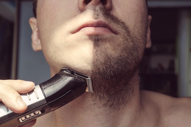 Jovem barbudo durante a preparação da barba usando aparador Metade do rosto com barba meio raspada