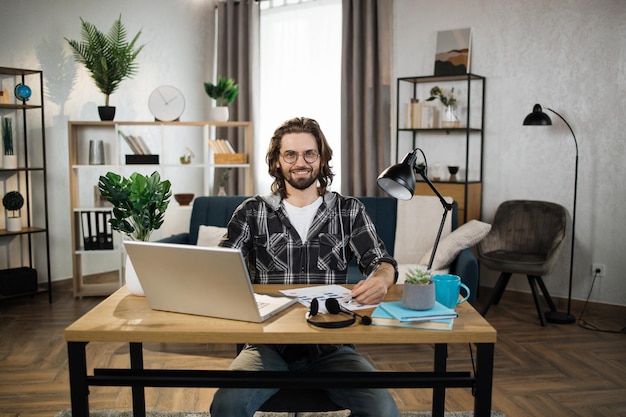 Foto jovem barbudo em camisa quadriculada e óculos sentado à mesa com laptop moderno