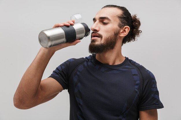 jovem barbudo em roupas esportivas bebendo água isolada sobre uma parede branca