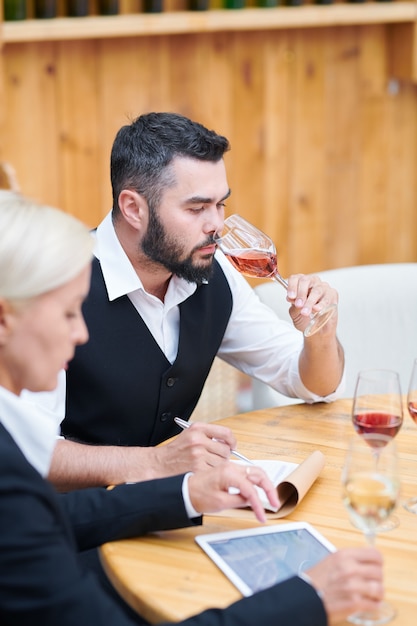 Jovem barbudo em trajes formais cheirando vários tipos de vinho enquanto faz anotações no bloco de notas perto do local de trabalho