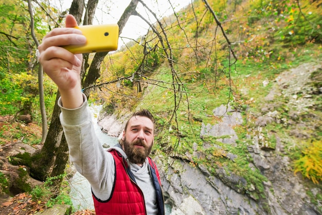 Jovem barbudo em um colete vermelho faz selfie em um telefone amarelo nas montanhas