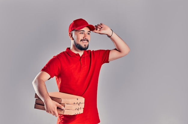 Jovem barbudo entregador de uniforme vermelho segura caixas de pizza isoladas em fundo cinza.