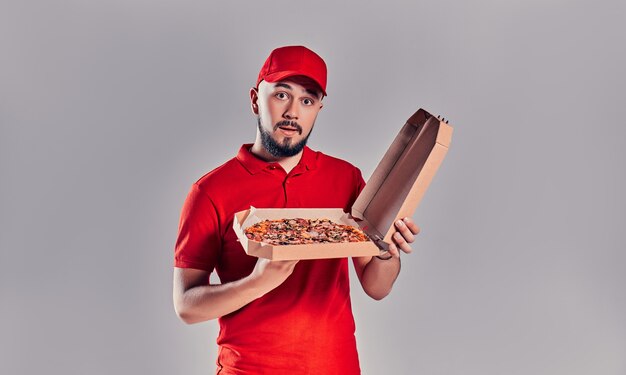 Jovem barbudo entregador de uniforme vermelho, segurando a caixa de pizza isolada no fundo cinza.