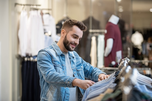 Jovem barbudo escolhendo roupas novas na prateleira de um shopping