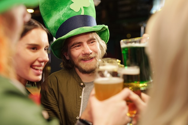 Jovem barbudo feliz com um chapéu tradicional irlandês segurando um copo de cerveja e olhando para um de seus amigos enquanto brinda pelo dia de São Patrício